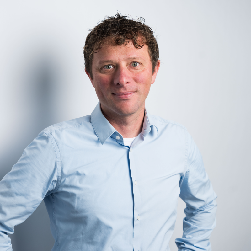 Smiling man in a light blue button-down shirt posing against a light gray background.